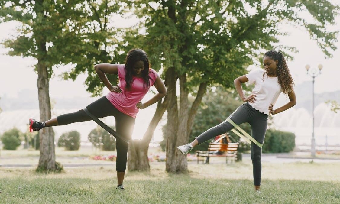 Two women exercising outdoors with resistance bands, focusing on lower body strength